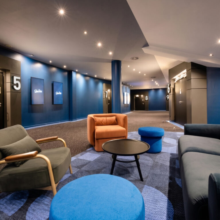 Cinema concourse lounge, breakout seating cluster, velvet upholstery, patterned modular carpet, recessed downlights, deep blue wall paint, wayfinding signage, auditorium entrance portal, commercial entertainment interior