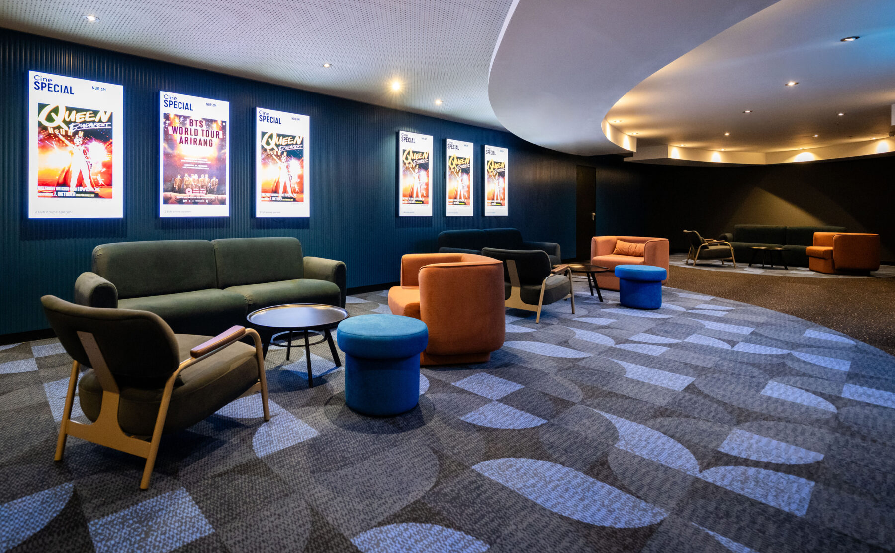 Cinema pre-function lounge, curved acoustic ceiling bulkhead, cove lighting, illuminated poster lightboxes, fluted wall cladding, geometric carpet tiles, modular tub chairs, velvet seating clusters, commercial cinema interior fit-out