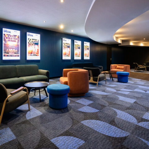 Cinema pre-function lounge, curved acoustic ceiling bulkhead, cove lighting, illuminated poster lightboxes, fluted wall cladding, geometric carpet tiles, modular tub chairs, velvet seating clusters, commercial cinema interior fit-out