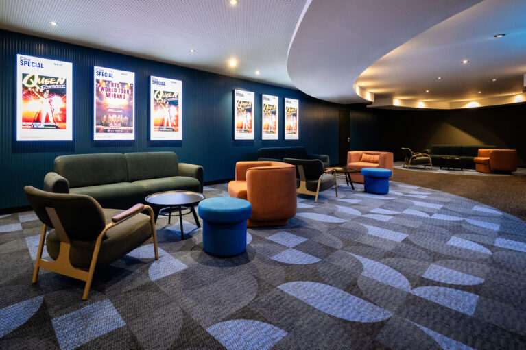 Cinema pre-function lounge, curved acoustic ceiling bulkhead, cove lighting, illuminated poster lightboxes, fluted wall cladding, geometric carpet tiles, modular tub chairs, velvet seating clusters, commercial cinema interior fit-out