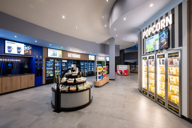 Self-service cinema concession, grab-and-go retail fit-out, illuminated popcorn warmers, beverage display refrigerators, fluted island counter, large format porcelain tiles, bulkhead downlighting, timber slat cabinetry, retail wayfinding signage
