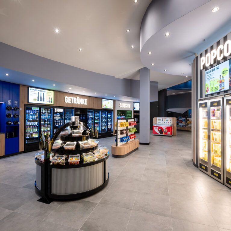 Self-service cinema concession, grab-and-go retail fit-out, illuminated popcorn warmers, beverage display refrigerators, fluted island counter, large format porcelain tiles, bulkhead downlighting, timber slat cabinetry, retail wayfinding signage
