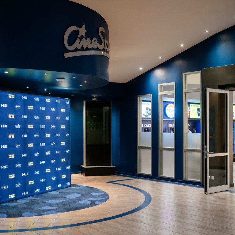 Step-and-repeat backdrop, curved architectural bulkhead, commercial entrance portal, interior glazing, porcelain floor tile, inset patterned carpet, recessed spotlighting, cinema foyer fit-out, retail marketplace entrance