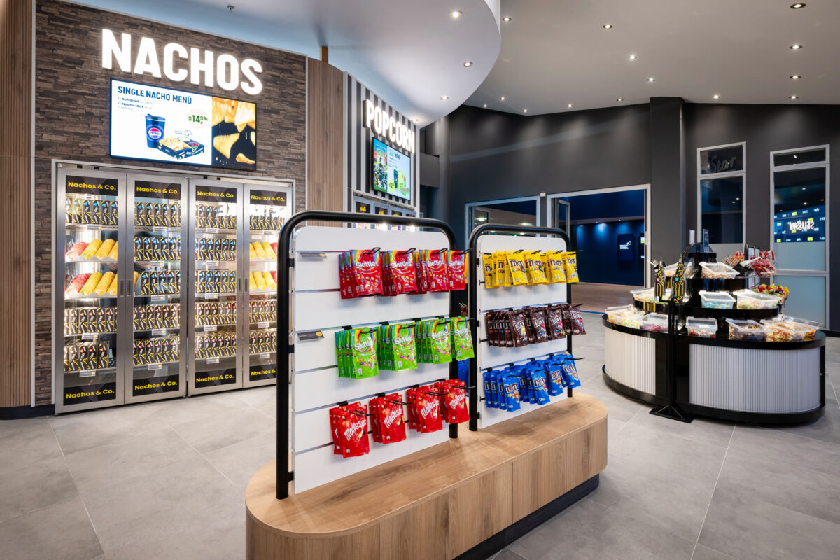 Retail merchandising gondola, tiered confectionery stand, heated nacho display case, stacked stone feature wall, illuminated channel letters, slatwall shelving unit, fluted display counter, commercial floor tile, grab-and-go cinema layout