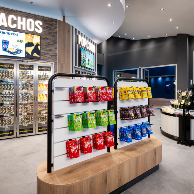 Retail merchandising gondola, tiered confectionery stand, heated nacho display case, stacked stone feature wall, illuminated channel letters, slatwall shelving unit, fluted display counter, commercial floor tile, grab-and-go cinema layout