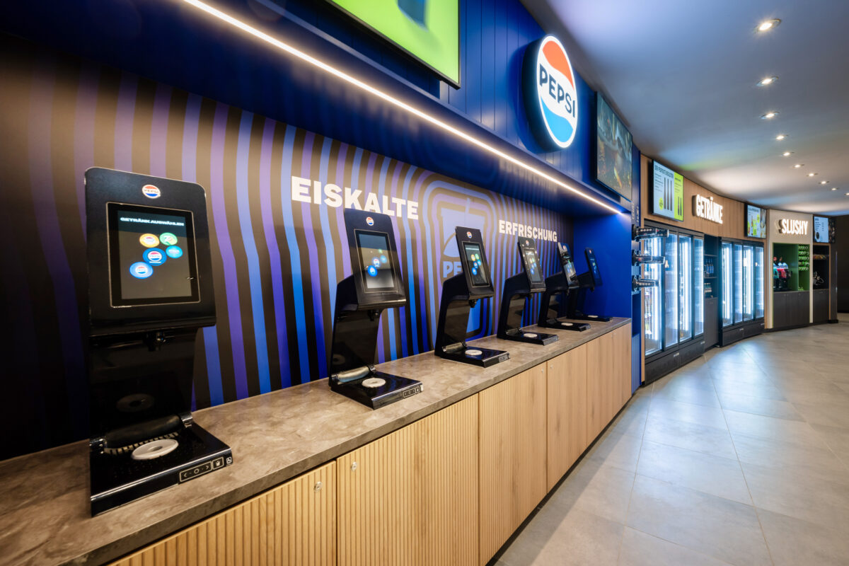 Automated soda fountain dispenser, touchscreen beverage station, stone composite countertop, vertical wood grain cabinetry, linear LED cove lighting, backlit brand signage, porcelain floor tile, self-service concession layout, cinema hospitality fit-out