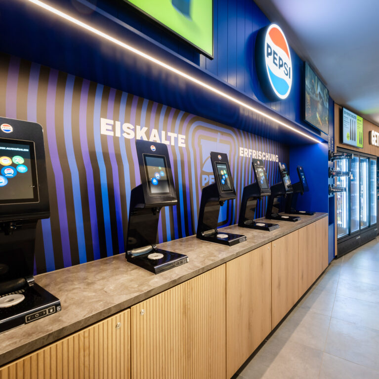 Automated soda fountain dispenser, touchscreen beverage station, stone composite countertop, vertical wood grain cabinetry, linear LED cove lighting, backlit brand signage, porcelain floor tile, self-service concession layout, cinema hospitality fit-out