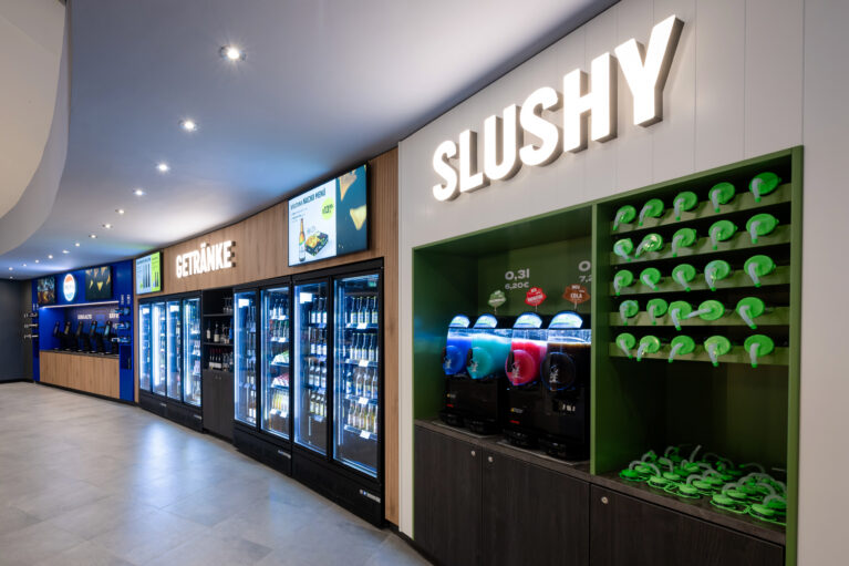Self-service beverage wall, slush machine station, custom cup display rack, glass door reach-in coolers, digital menu monitors, illuminated channel lettering, vertical wall paneling, retail joinery fit-out, recessed ceiling spots
