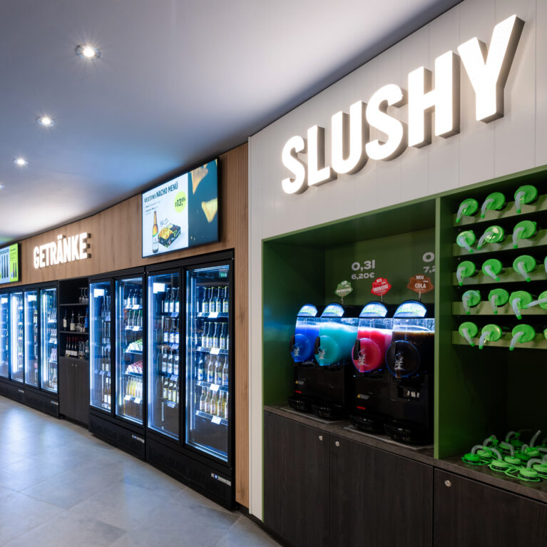 Self-service beverage wall, slush machine station, custom cup display rack, glass door reach-in coolers, digital menu monitors, illuminated channel lettering, vertical wall paneling, retail joinery fit-out, recessed ceiling spots