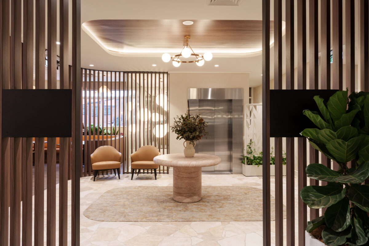 Travertine pedestal table, timber batten screens, elevator lobby design, brass ring chandelier, recessed ceiling cove, crazy paving floor, textured area rug, cognac leather armchairs, indoor greenery, commercial foyer fit-out