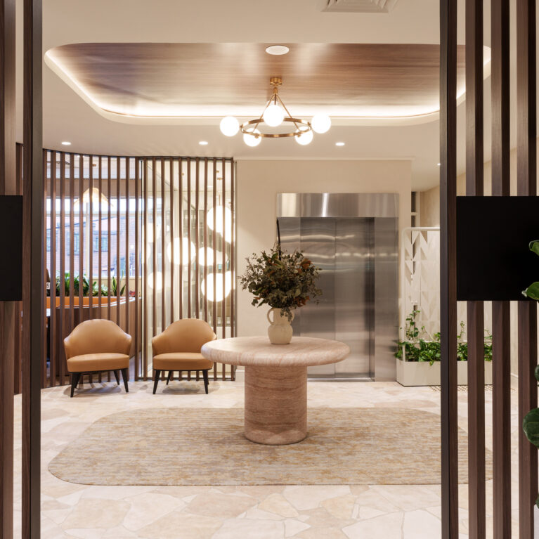 Travertine pedestal table, timber batten screens, elevator lobby design, brass ring chandelier, recessed ceiling cove, crazy paving floor, textured area rug, cognac leather armchairs, indoor greenery, commercial foyer fit-out