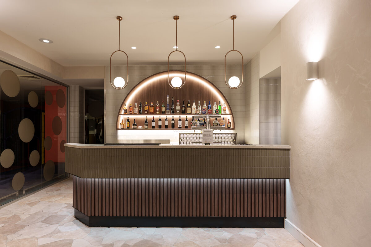 Fluted timber apron, olive kit-kat tile counter, copper oval pendants, arched back bar, under-counter LED strip, crazy paving stone floor, cylindrical wall sconce, textured plaster finish, glass partition wall, hospitality bar design