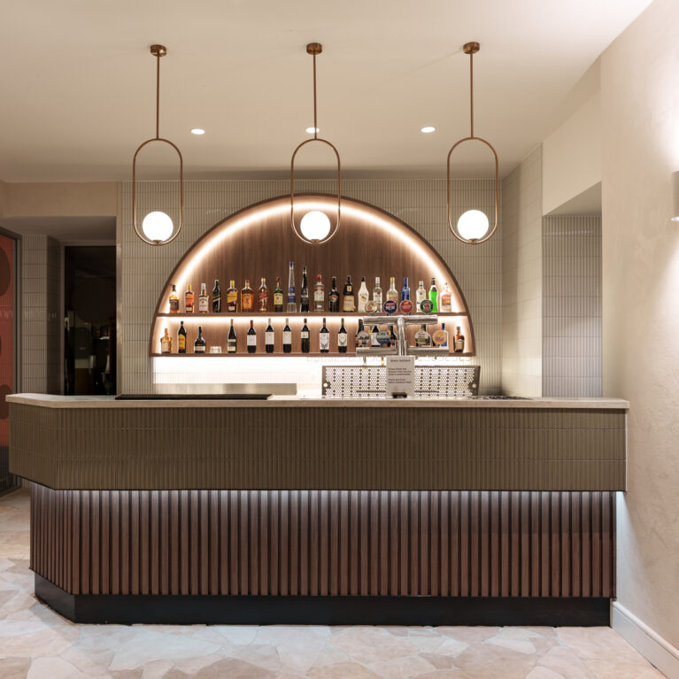 Fluted timber apron, olive kit-kat tile counter, copper oval pendants, arched back bar, under-counter LED strip, crazy paving stone floor, cylindrical wall sconce, textured plaster finish, glass partition wall, hospitality bar design