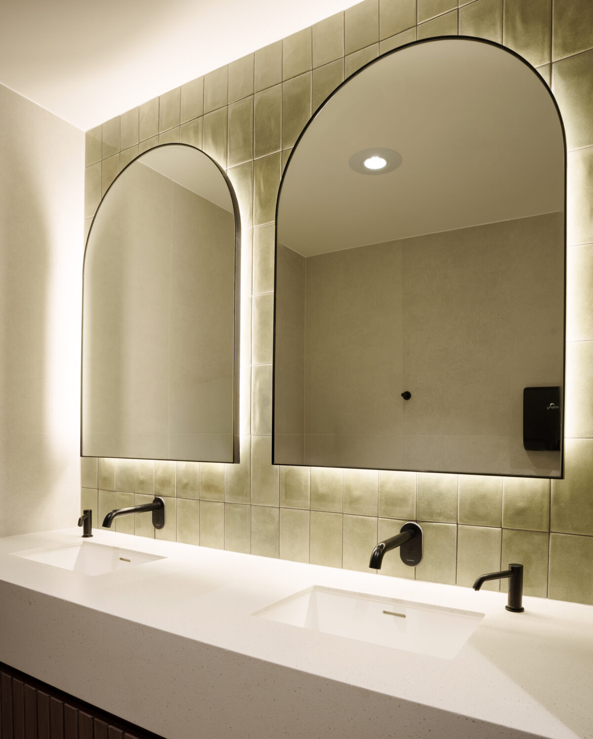 Arched LED mirrors, olive green glazed tiles, zellige ceramic wall tiles, matte black tapware, wall-mounted spouts, undermount basins, engineered stone vanity, backlighting, commercial washroom design, club amenities