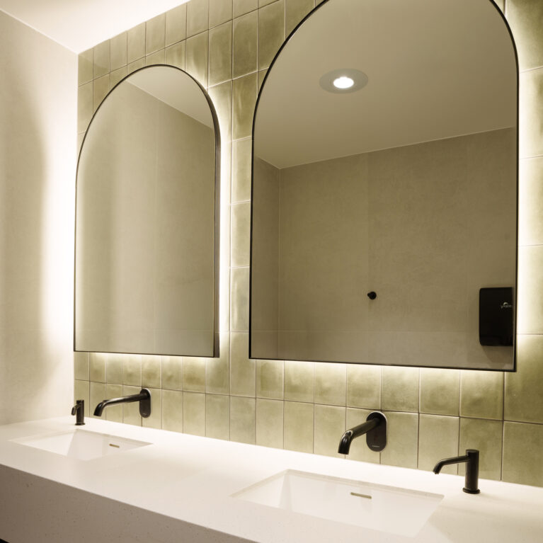 Arched LED mirrors, olive green glazed tiles, zellige ceramic wall tiles, matte black tapware, wall-mounted spouts, undermount basins, engineered stone vanity, backlighting, commercial washroom design, club amenities