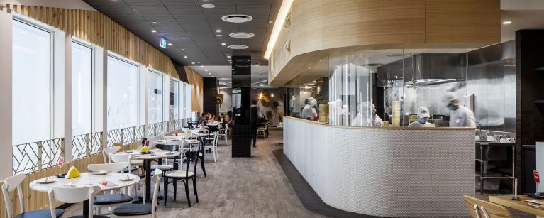 A commercial restaurant design highlighting an open show kitchen with glass panels, curved timber bulkheads, linear window seating tables, and textured grey floor tiling.