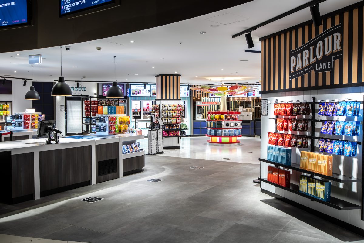 A modern cinema candy bar renovation featuring a dark laminate service counter, timber-slat wall paneling, industrial black pendant lights, and modular concession display racks.