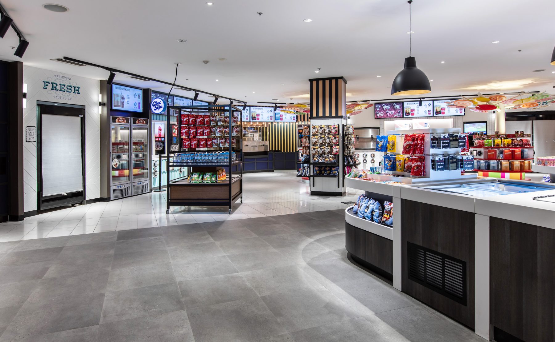 Commercial floor transition, open-plan cinema foyer, dark stone tiles, freestanding retail gondolas, matte black pendant lighting, black track spotlights, architectural zoning, integrated service joinery.