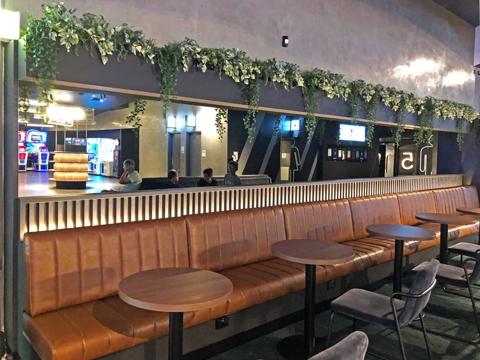 A modern lobby banquette with tan cushioned seating, dark tables, and a mirror reflecting the candy bar.