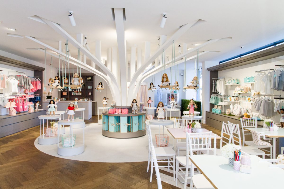 Boutique retail showroom, sculptural tree feature, miniature hanging swings, parquet timber flooring, white wooden cafe tables, custom doll display stands, integrated lifestyle zoning.