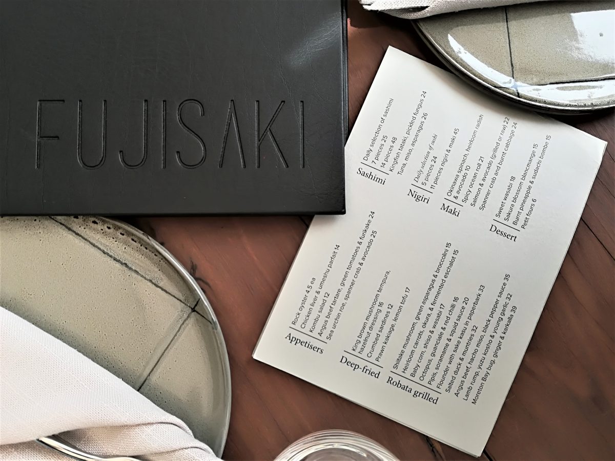 A luxury Japanese restaurant tabletop presentation featuring a debossed black leather menu booklet, printed cream course menus, artisanal ceramic plates, and natural linen napkins.