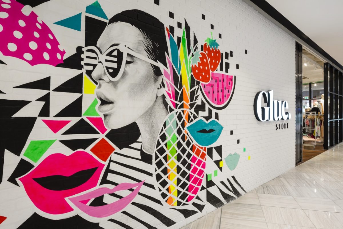 Street art retail storefront, pop art graphic mural, black dimensional logo, white brick wall finish, glazed entry door frame, marble concourse flooring, mall shop facade.