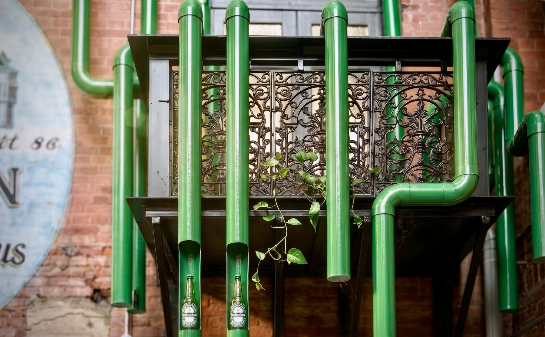 Hospitality brand activation, glossy green pipes, industrial pipe sculpture, wrought iron balcony, historic brick wall, integrated product display, LED bottle illumination.