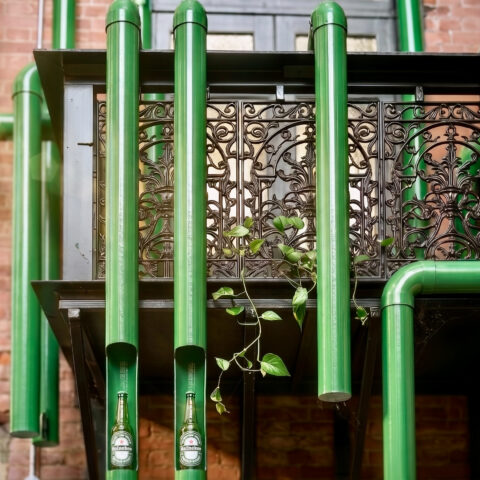 Hospitality brand activation, glossy green pipes, industrial pipe sculpture, wrought iron balcony, historic brick wall, integrated product display, LED bottle illumination.