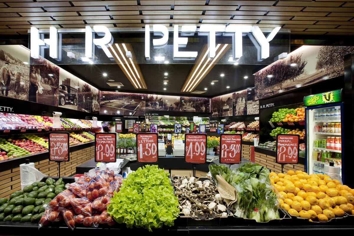 Illuminated retail signage, timber ceiling baffles, fresh market layout, tiered produce displays, monochrome wall panels, linear strip lighting, commercial food refrigeration.