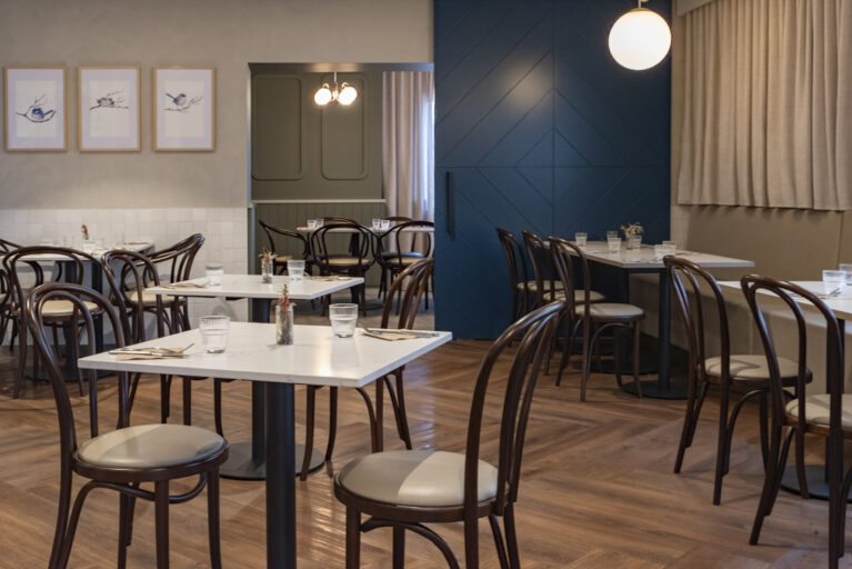 A calm and contemporary bistro space defined by chevron timber flooring, classic bentwood chairs and crisp stone-look tabletops. Gentle colour blocking, soft drapery and warm lighting create an inviting dining room that balances familiarity with a refined, modern edge.