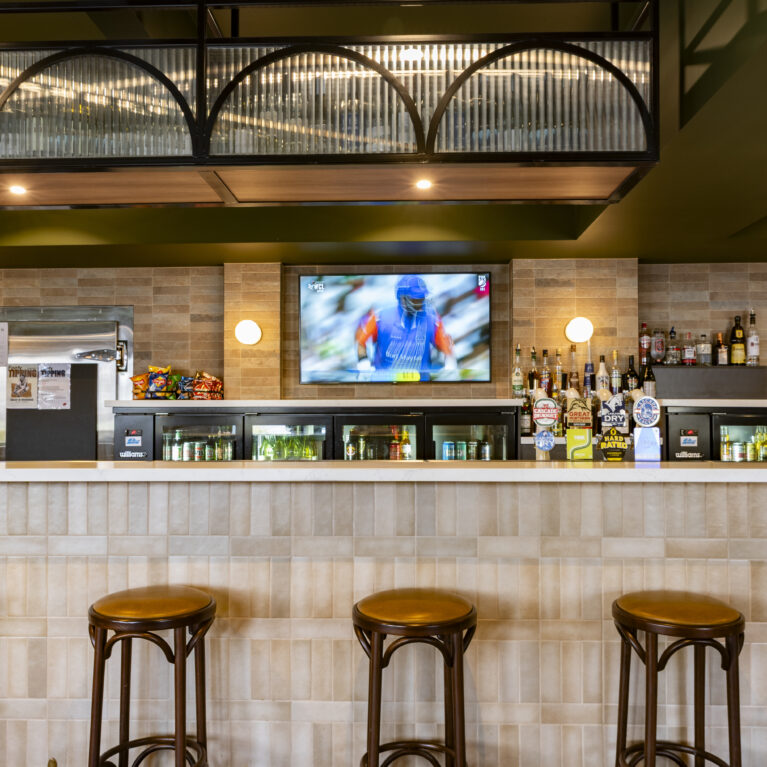 A warm, tactile composition of Safi clay tiles, engineered stone, and dark oak stools, anchored by a softly illuminated bar display.