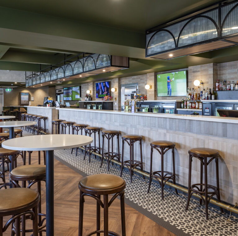 A lively, contemporary sports bar with warm timber tones, patterned flooring and a long, tiled bar front. Classic bentwood stools, robust materials and layered lighting create a relaxed, social atmosphere built for all-day dining, casual drinks and high-energy match nights.