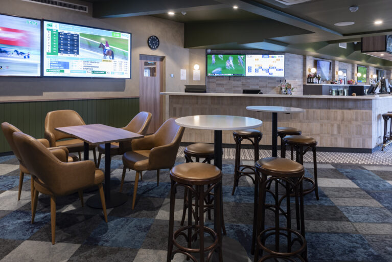 A relaxed sports lounge combining upholstered comfort, classic timber stools and a bold patterned carpet. Layered materials, muted colours and multiple screens shape an easygoing environment designed for long stays, casual gatherings and an uninterrupted view of the action.
