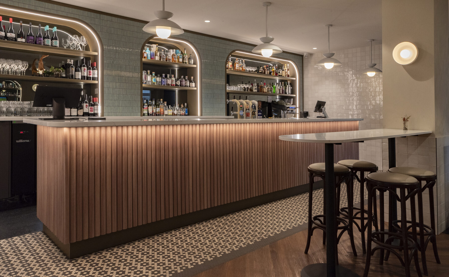 A refined yet approachable bar anchored by fluted timber, softly lustred tiles and arched illuminated shelves. Warm lighting, classic bentwood stools and patterned mosaic flooring create a contemporary hospitality setting with an unmistakable Tasmanian pub soul.
