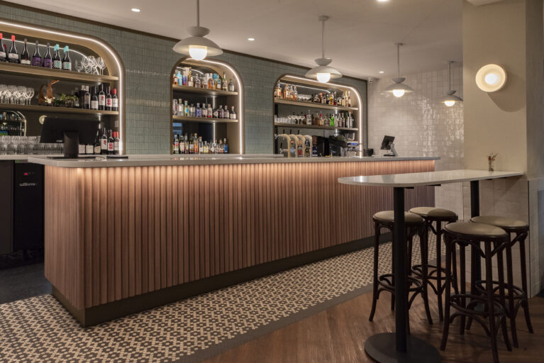 A refined yet approachable bar anchored by fluted timber, softly lustred tiles and arched illuminated shelves. Warm lighting, classic bentwood stools and patterned mosaic flooring create a contemporary hospitality setting with an unmistakable Tasmanian pub soul.