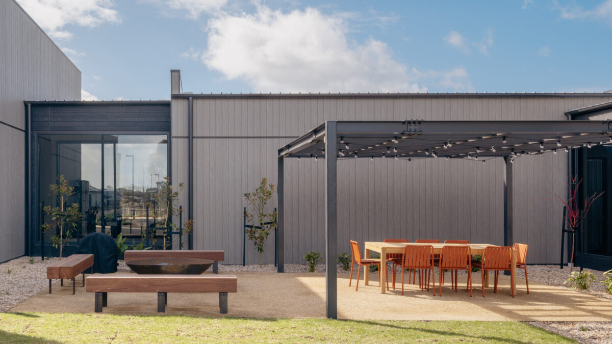 A robust pergola shelters an outdoor table setting, paired with timber-and-steel benches around a fire bowl. The space hosts everything from long lunches to evening events, connecting directly to garden paths and planting.