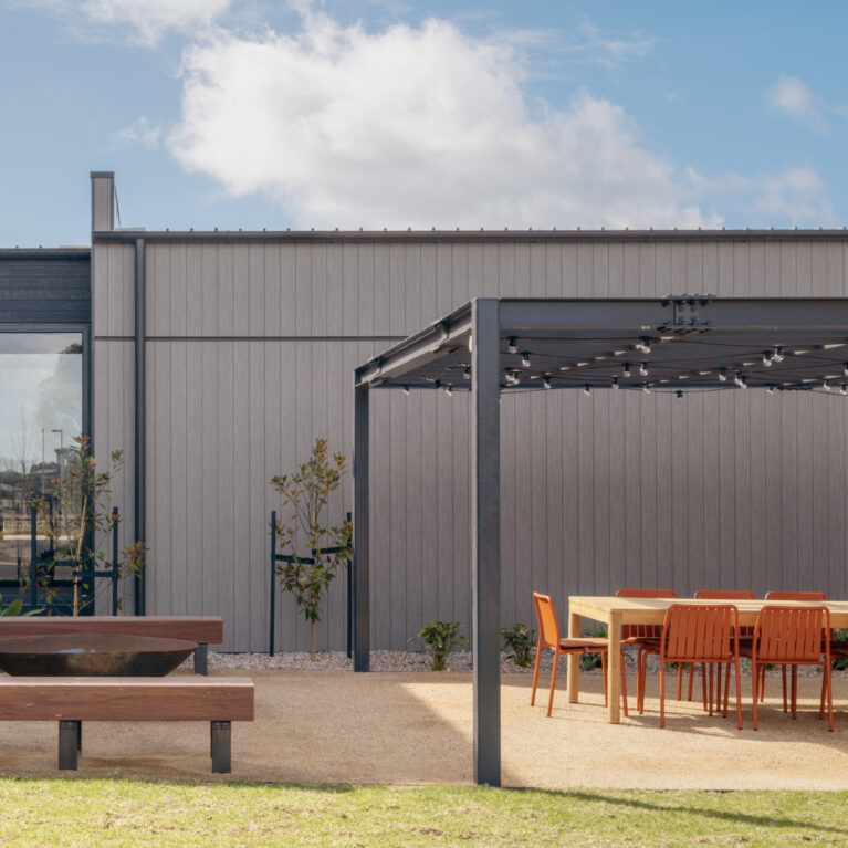 A robust pergola shelters an outdoor table setting, paired with timber-and-steel benches around a fire bowl. The space hosts everything from long lunches to evening events, connecting directly to garden paths and planting.