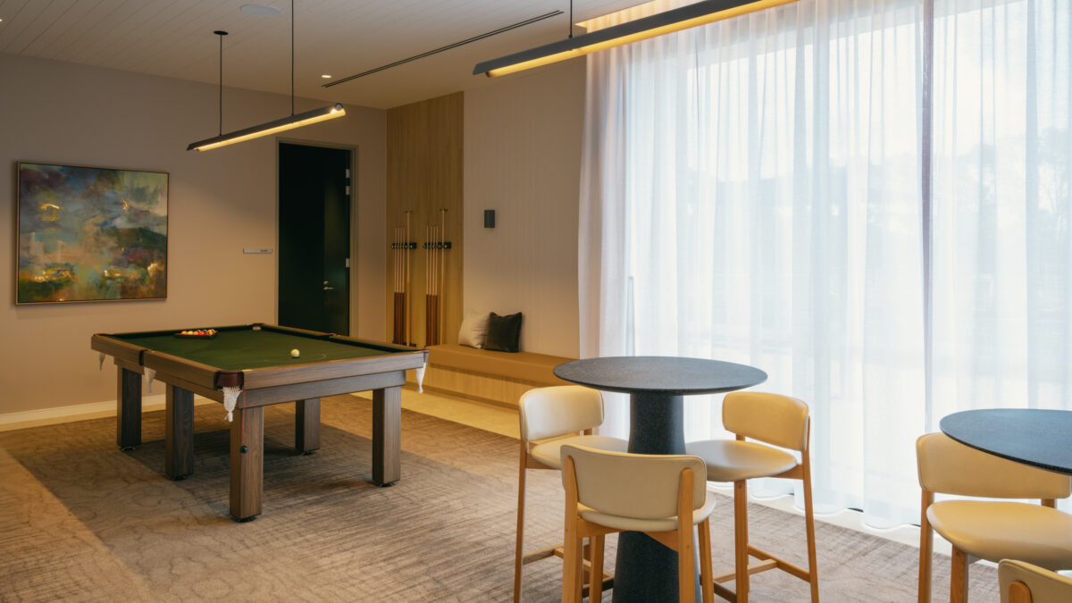 A club-style games lounge pairs a full-size billiards table with café-height tables beside sheer curtains. Upholstered bench seating, long linear pendants and acoustic treatment temper the buzz, encouraging everything from tournaments to quiet card games and morning coffees.