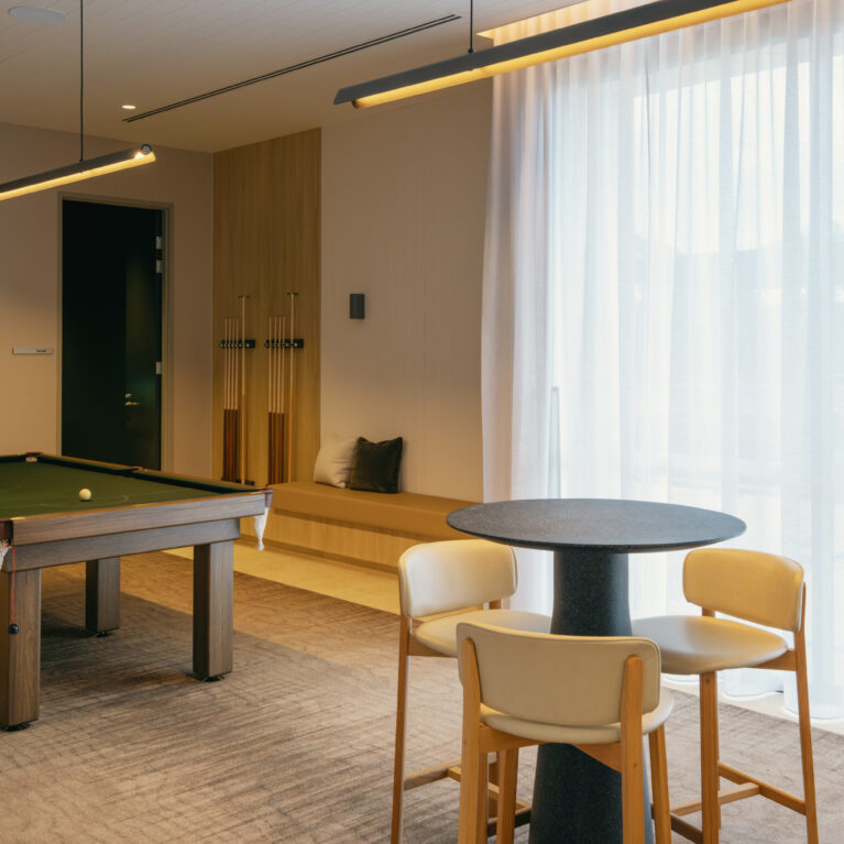 A club-style games lounge pairs a full-size billiards table with café-height tables beside sheer curtains. Upholstered bench seating, long linear pendants and acoustic treatment temper the buzz, encouraging everything from tournaments to quiet card games and morning coffees.