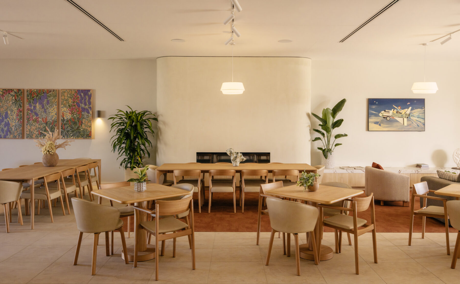 A light-filled dining room anchored by a soft, curved fireplace. Timber tables, upholstered chairs and artful planting create the feeling of an all-day neighbourhood café.
