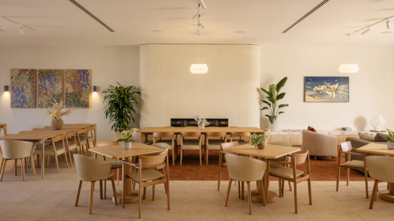 A light-filled dining room anchored by a soft, curved fireplace. Timber tables, upholstered chairs and artful planting create the feeling of an all-day neighbourhood café.