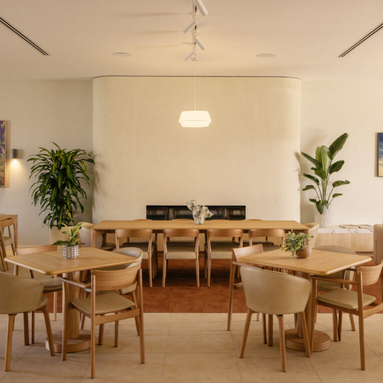 A light-filled dining room anchored by a soft, curved fireplace. Timber tables, upholstered chairs and artful planting create the feeling of an all-day neighbourhood café.