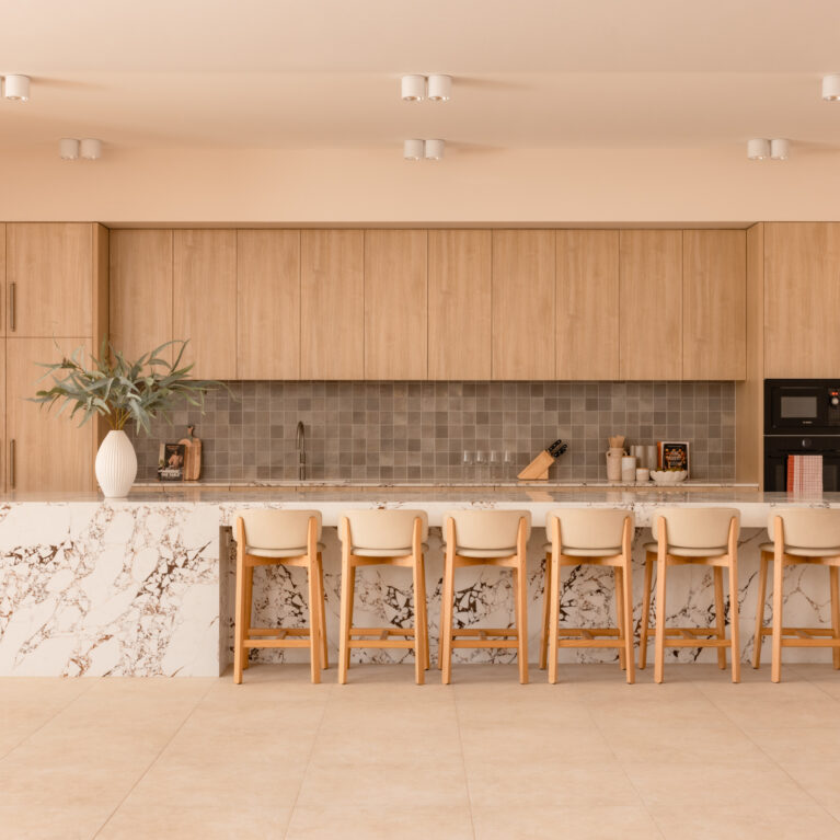 A generous island anchors the residents’ kitchen, pairing a boldly veined stone with pale timber joinery. Soft task lighting washes a square-tiled splashback, while integrated appliances and slimline stools turn the space into a relaxed hub for cooking, classes and communal dining.