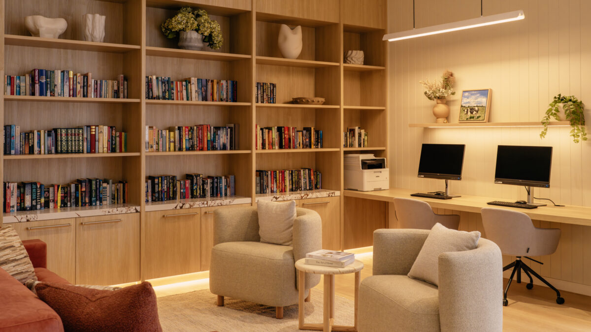 A companion view of the library shows the dedicated study bench with two screens, cable management and soft task lighting. Open shelving continues overhead for display, while a pair of sculptural armchairs create a secondary conversation pocket in the foreground.
