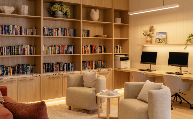 A companion view of the library shows the dedicated study bench with two screens, cable management and soft task lighting. Open shelving continues overhead for display, while a pair of sculptural armchairs create a secondary conversation pocket in the foreground.