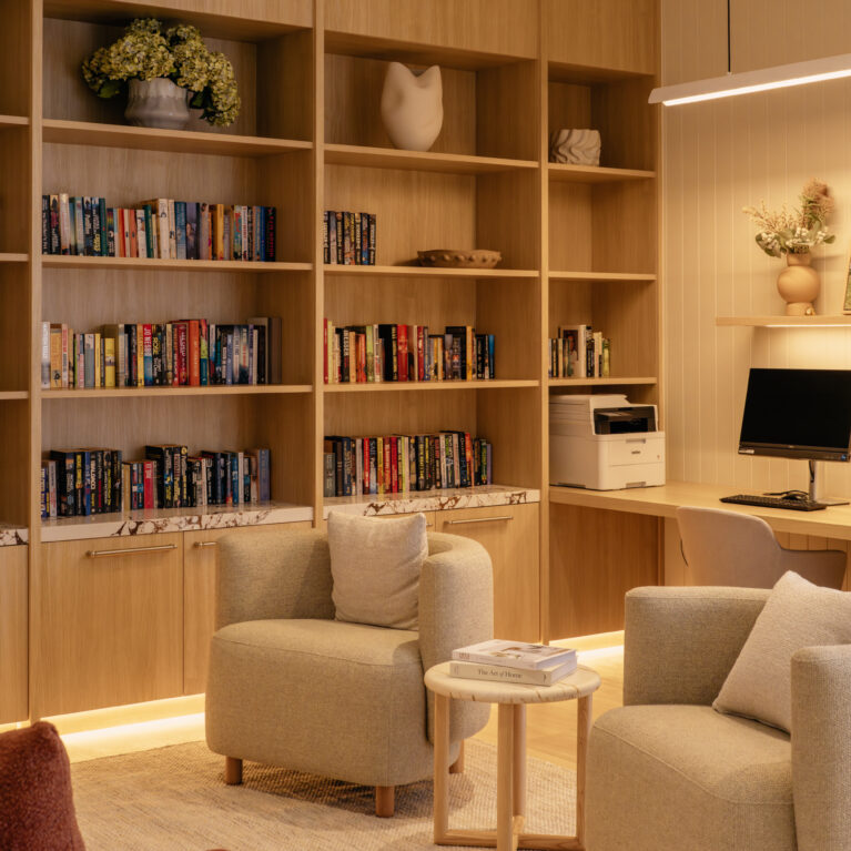 A companion view of the library shows the dedicated study bench with two screens, cable management and soft task lighting. Open shelving continues overhead for display, while a pair of sculptural armchairs create a secondary conversation pocket in the foreground.