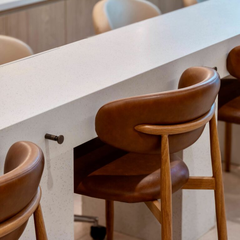 This close-up captures the manicure station seating, combining tactile richness and functionality. Soft leather chairs with wooden frames pair seamlessly with the crisp, solid surface table, while discreet hooks offer clients a practical spot to hang personal belongings.