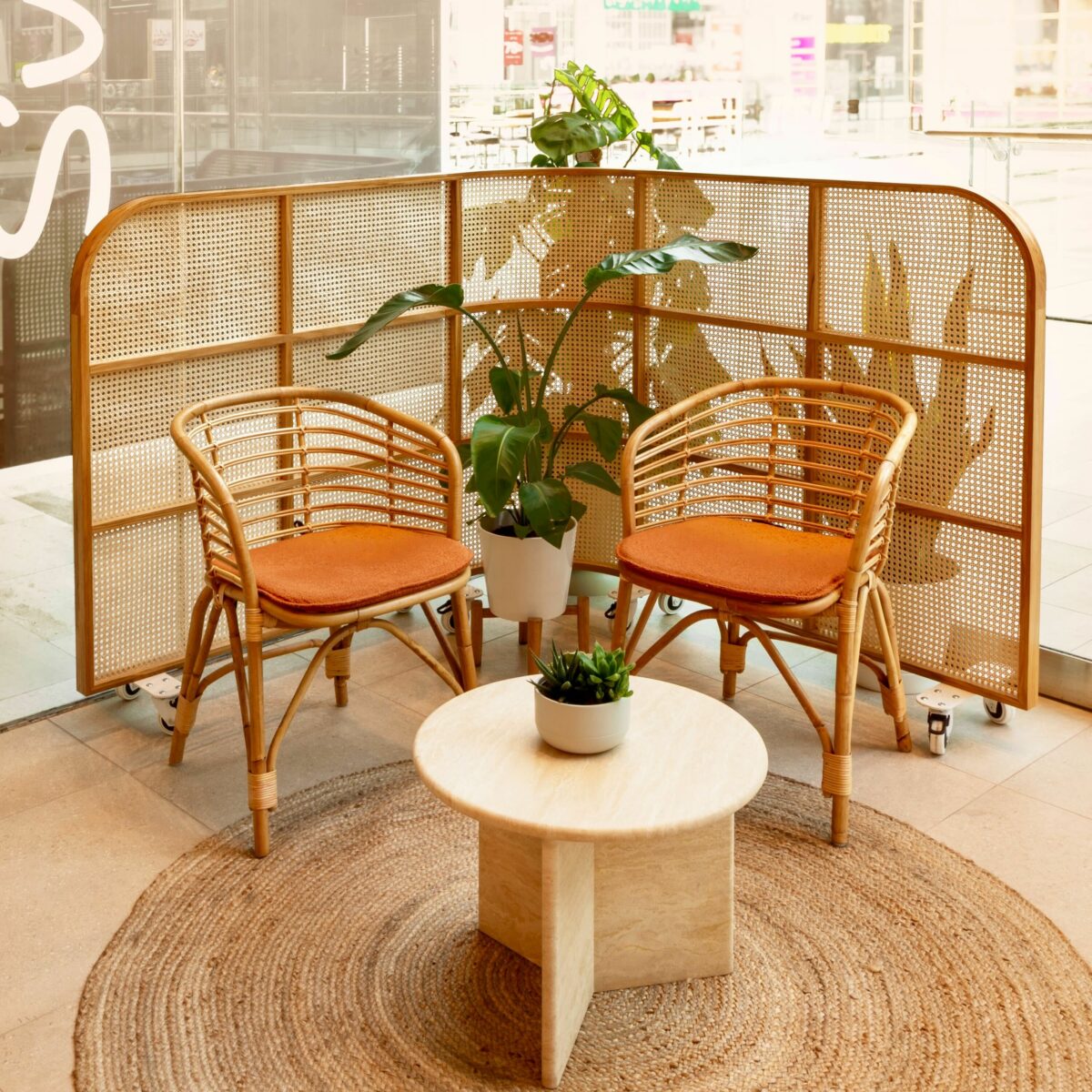The waiting area creates a cozy, welcoming corner with rattan chairs, earthy-toned cushions, and a woven rug, complemented by potted greenery. A compact travertine coffee table completes the setting, blending natural textures with a warm, modern aesthetic.