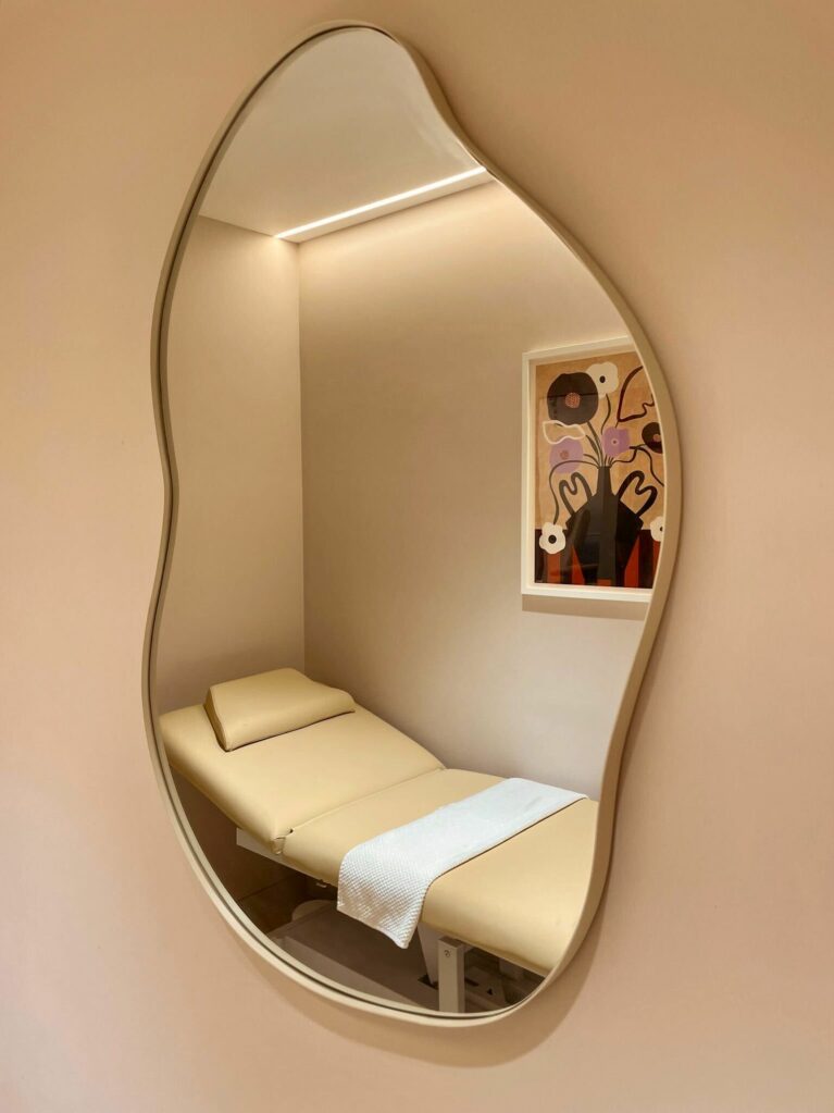This treatment room evokes serenity with its soft, neutral palette and calming textures. The organic mirror shape adds playfulness, while indirect lighting enhances the soothing ambiance, creating an intimate environment for personalized beauty treatments.