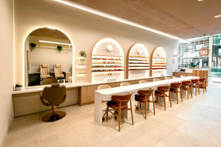 A bright, airy workspace where clients enjoy nail and threading services in comfort, framed by softly lit arches showcasing a curated polish display.